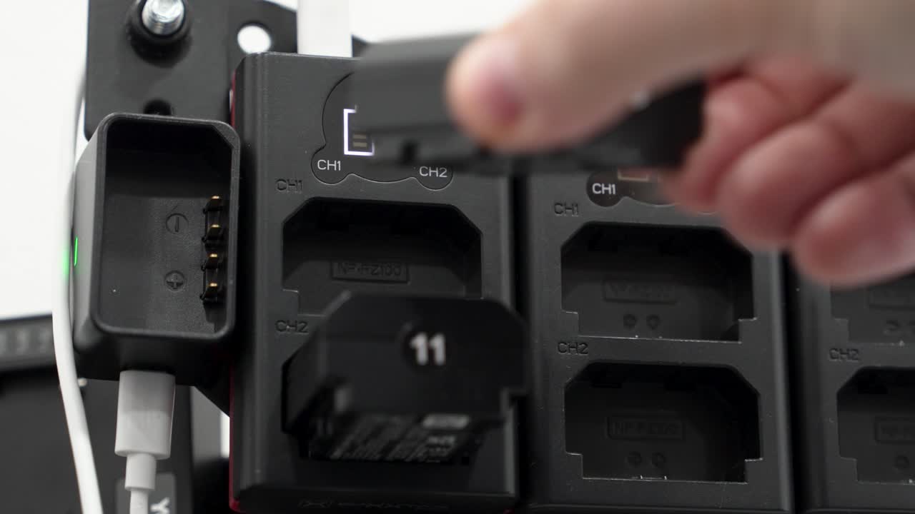 A person uses a battery charging dock to charge two batteries. The dock has lights that show the charging status. This happens in a well-lit workshop during the day