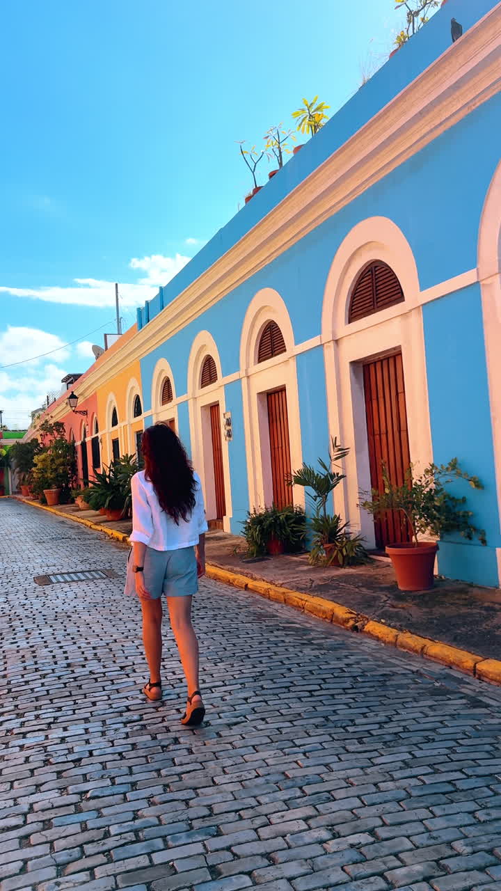 Donna Bruna con Capelli Lunghi che Cammina Lungo il Vicolo Pavimentato Accanto alla Casa Colorata. Bellissimi Vasi di Fiori sono Vicino agli Edifici. Porto Rico. Video Verticale