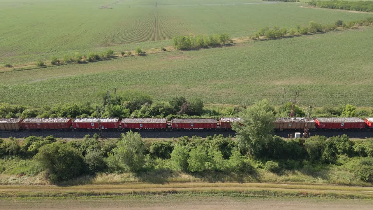 A freight train travels along railway tracks cutting through a vast green agricultural field. The aerial view captures the contrast between industrial transport and the surrounding rural environment.