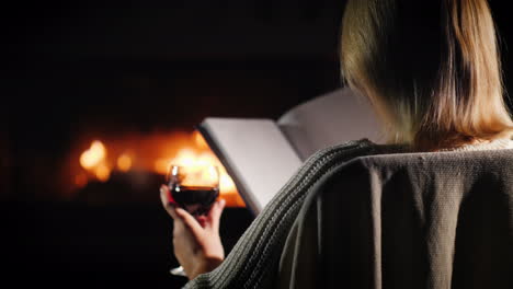 mujer con un libro y una copa de vino se relaja en casa junto a la chimenea