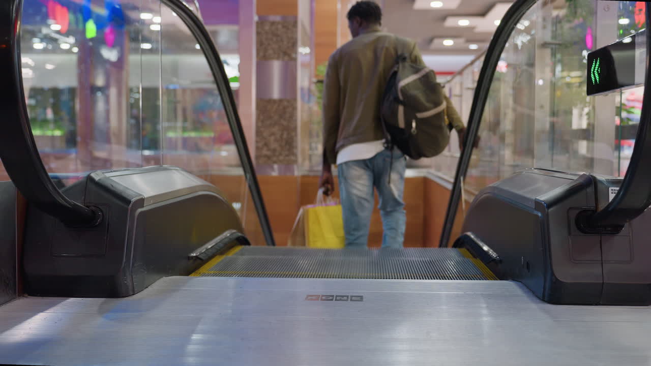 Back view of shopper wearing sneakers and blue jeans holding yellow and brown shopping bags stepping off descending escalator in modern shopping mall with lights, glass railing