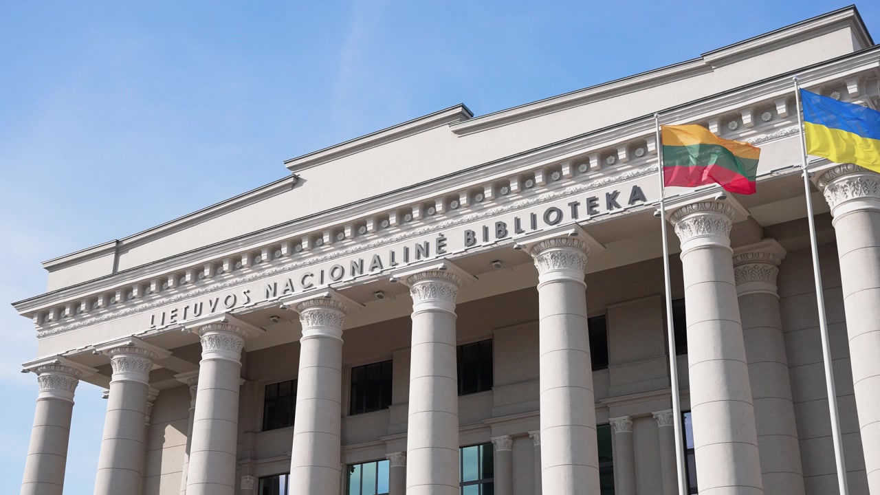 Stately columns and classic architecture of the Martynas Mažvydas National Library under clear blue sky, with Lithuanian and Ukrainian flags waving. Wide angle, daylight, nobody visible.