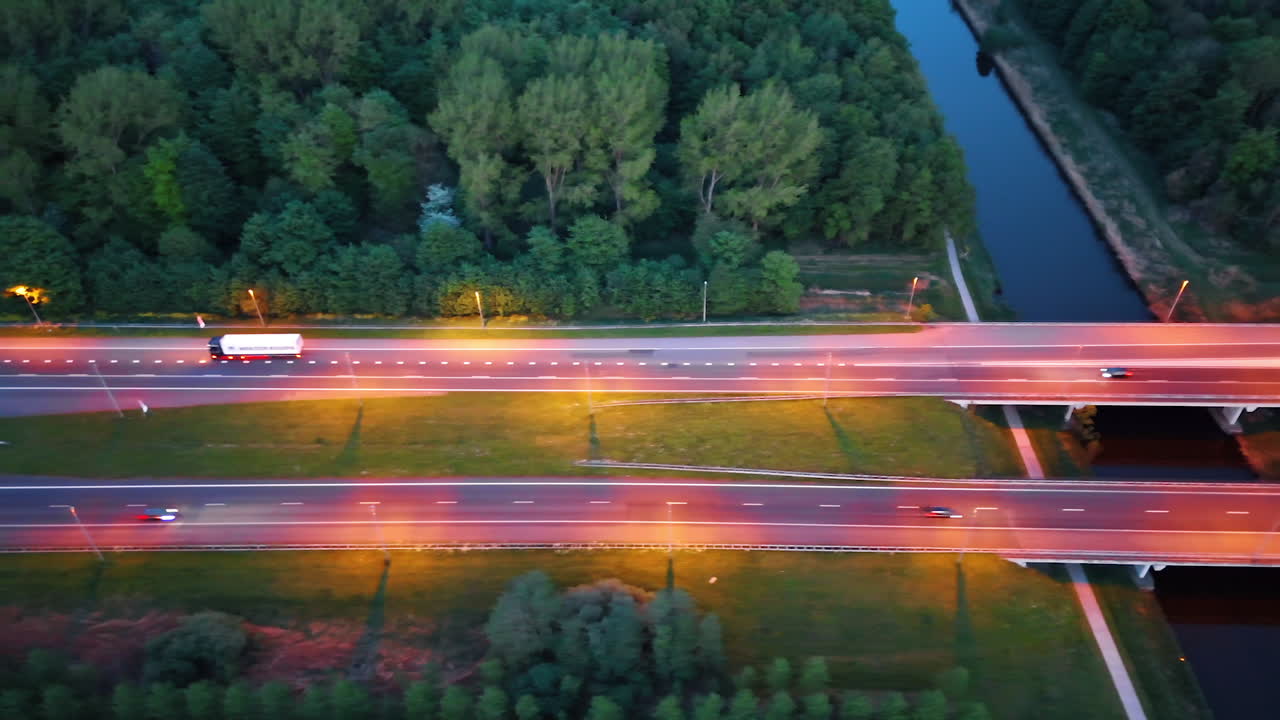 Cars move by the roads illuminated pink at dusk time. Flight over the canal and forest. The Netherlands.