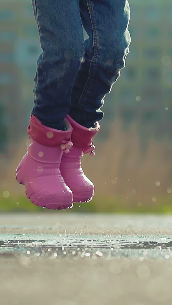 CU, slow motion: funny little girl wearing cute pink rubber boots and jeans has fun jumping in large puddle in sunny spring park after rain closeup