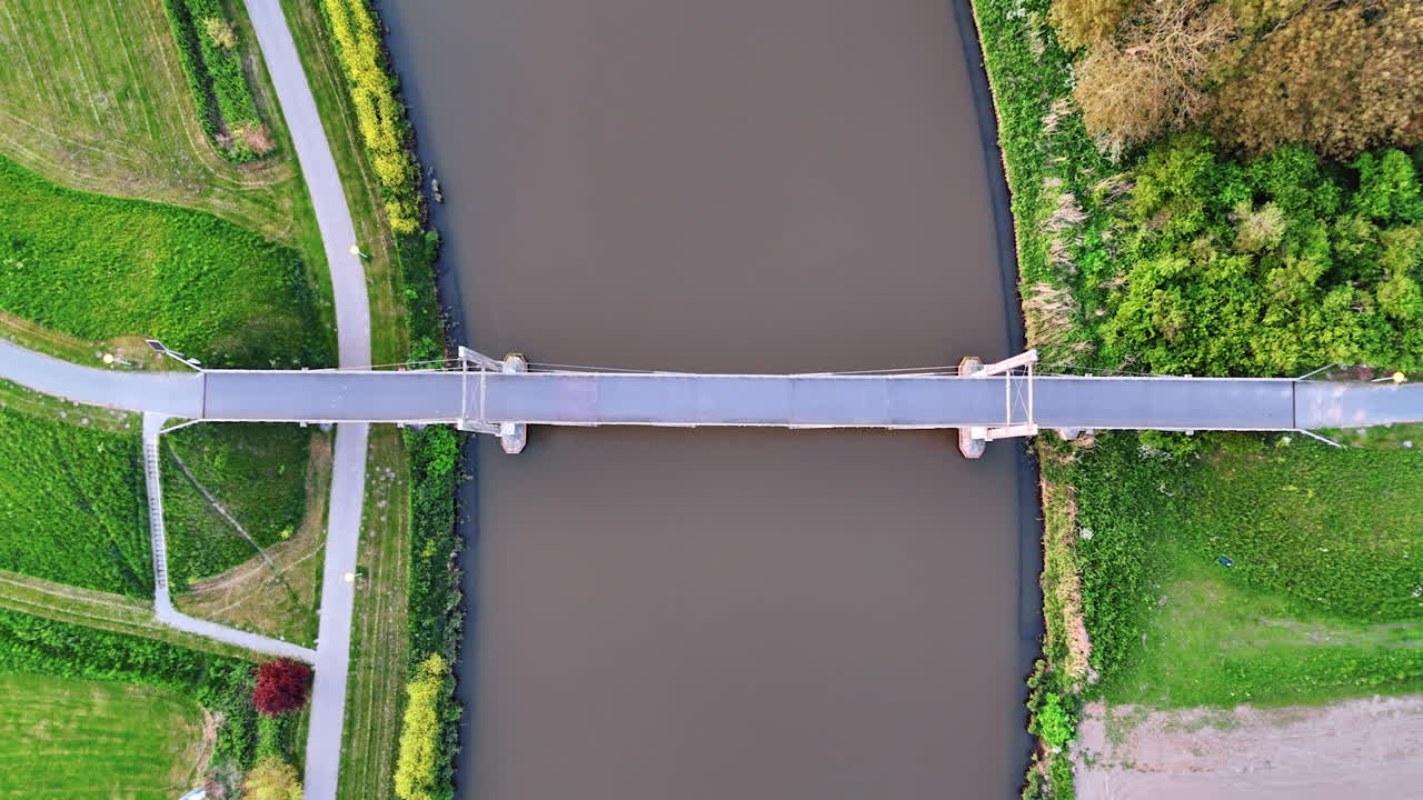 Suspension bridge over the narrow river. Beautiful green waterfronts along the waterscape. Top view.