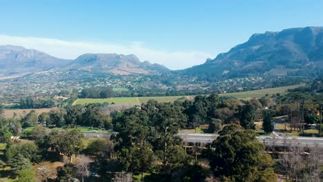 Hovering-above-a-park-in-Cape-Town,-South-Africa-overlooking-a-vista-of-mountains,-greenery-and-highways