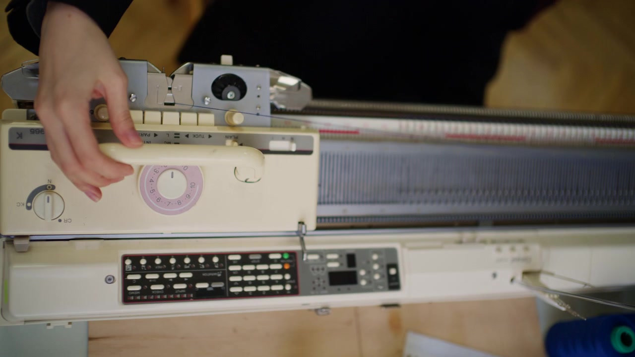 Primo piano di una macchina per maglieria con aghi metallici, guida di misurazione numerata, carrello e sistema di tensione del filo.