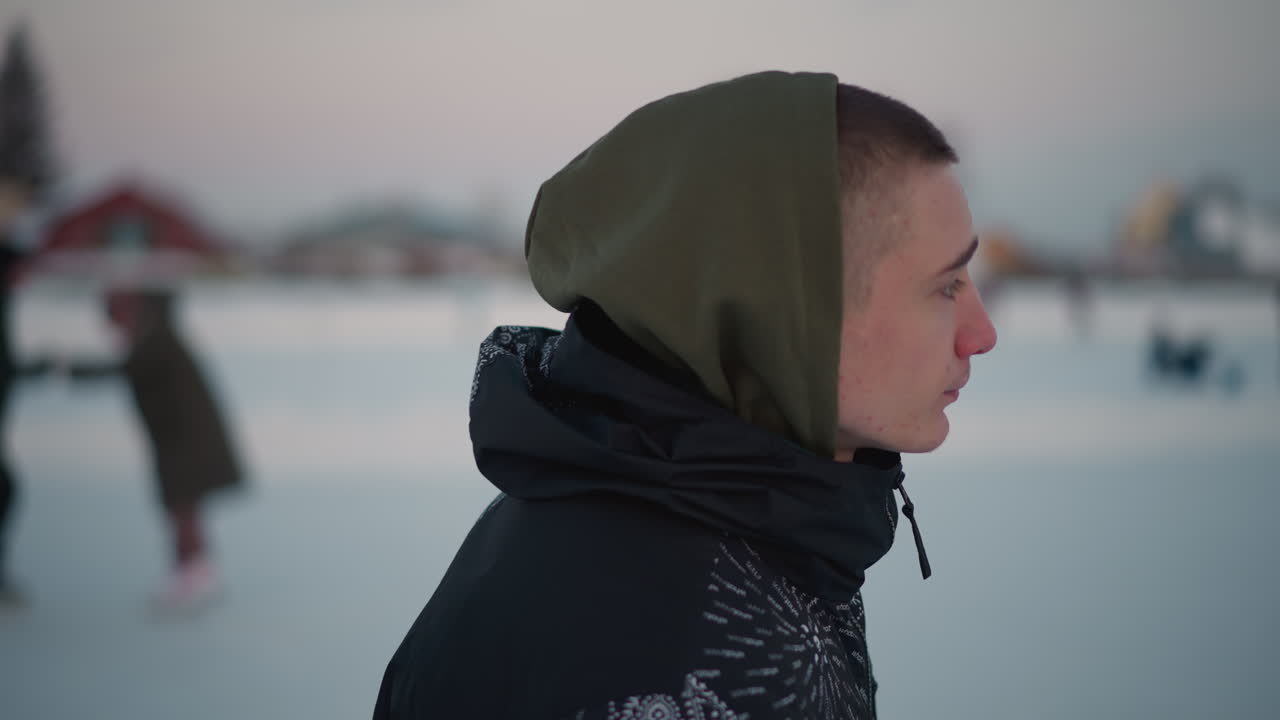 young man skating on ice rink wearing patterned jacket and hoodie, blurred background of other skaters, snow covered ice with blade marks under pale winter sky near bare trees, distant buildings