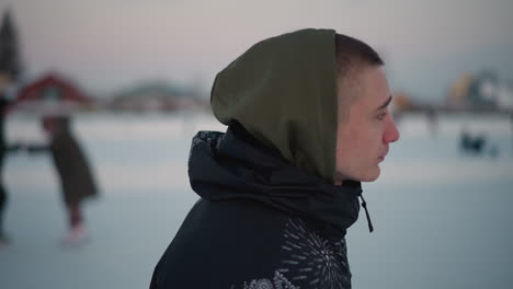 young man skating on ice rink wearing patterned jacket and hoodie, blurred background of other skaters, snow covered ice with blade marks under pale winter sky near bare trees, distant buildings