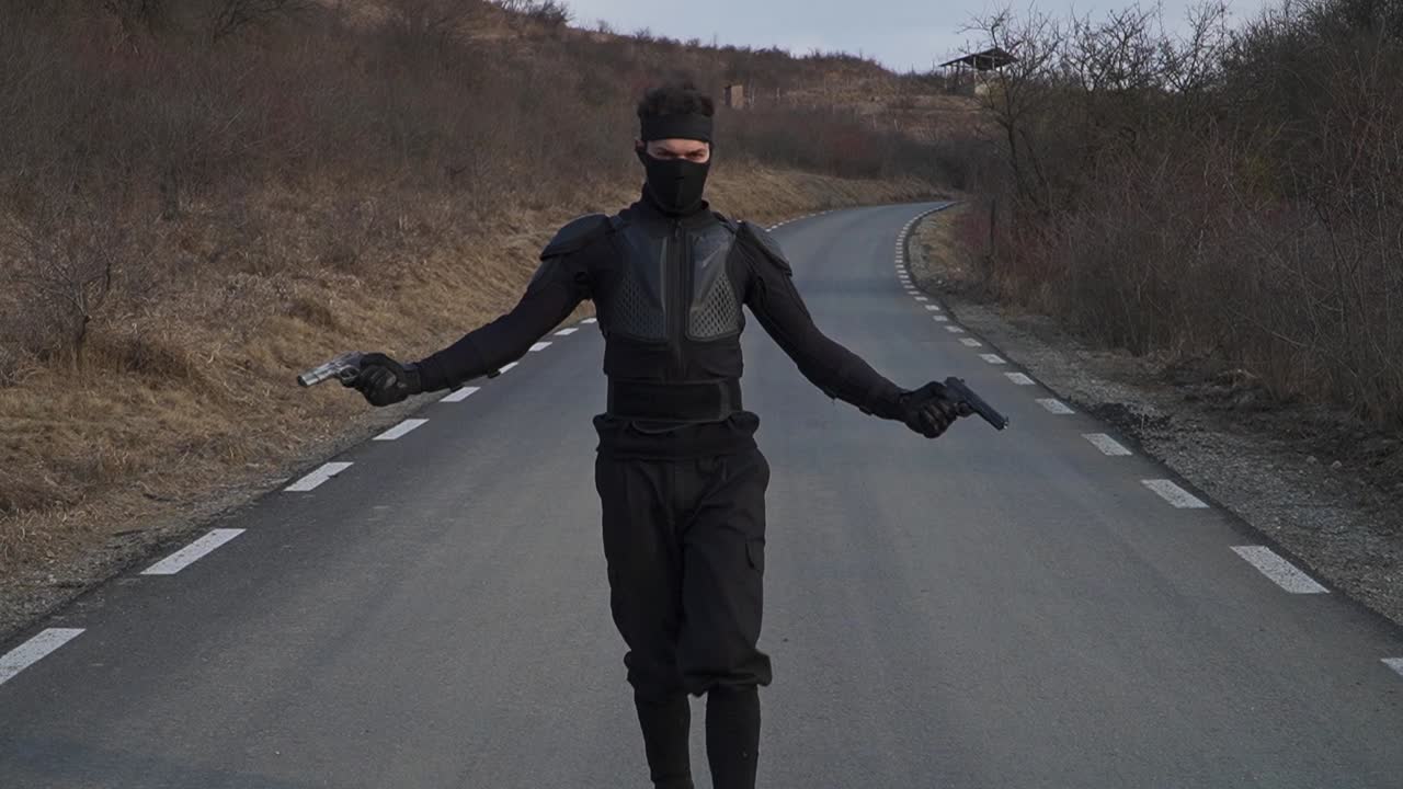 Cinematic scene of a man dressed in black, partially masked and armed with two pistols, walking with a confident and intense attitude down a lonely road.