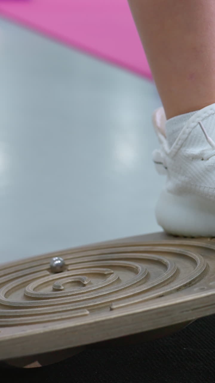 핑크색 옷을 입은 어린 소녀가 색 운동화로 런스 보드에서 균형을 잡으면서 아래로 초점을 맞추고, 다른 아이들이 운동을 할 때 집중력으로 자세를 조정합니다.