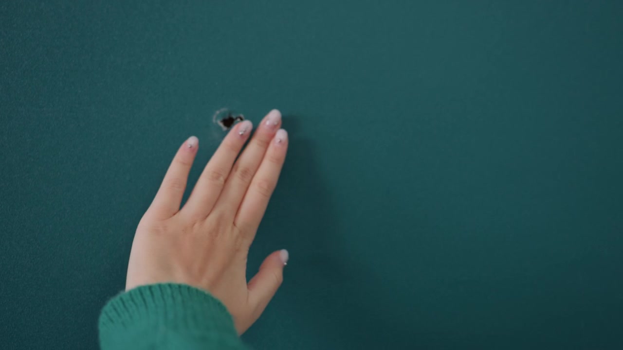 Giovane donna caucasica ispeziona un piccolo buco nella parete color verde acqua dell'appartamento, le punte delle dita che tracciano l'intonaco ruvido mentre lo sguardo di profilo studia il danno. L'inquilina si ferma durante il trasloco, l'appassionata di fai da te immagina un piano di riparazione.