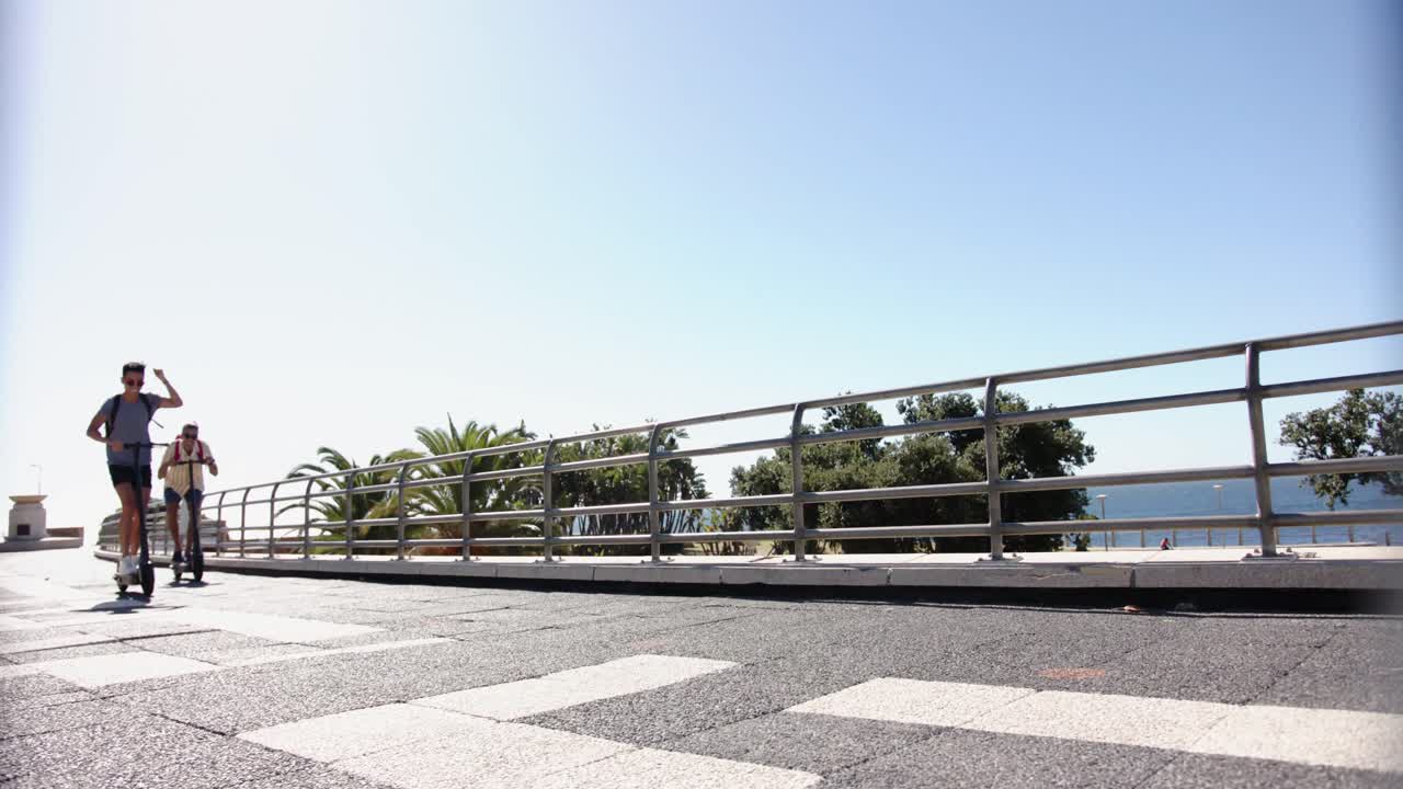 Heureux couple gay mixte utilisant des trottinettes sur la promenade en bord de mer, au ralenti.