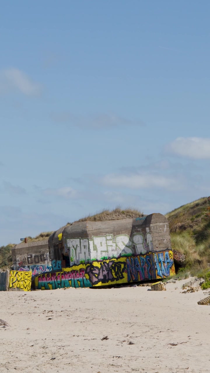 La telecamera panoramica su una spiaggia sabbiosa, rivelando bunker storici con graffiti sotto una luce diurna brillante, paesaggio costiero