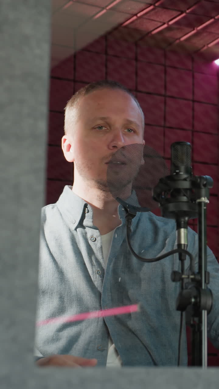 un hombre con una camisa de manga larga azul se para frente a un micrófono de estudio, sosteniendo un auricular cerca de su oreja en una habitación a prueba de sonido roja. la escena captura un momento de escucha cuidadosa