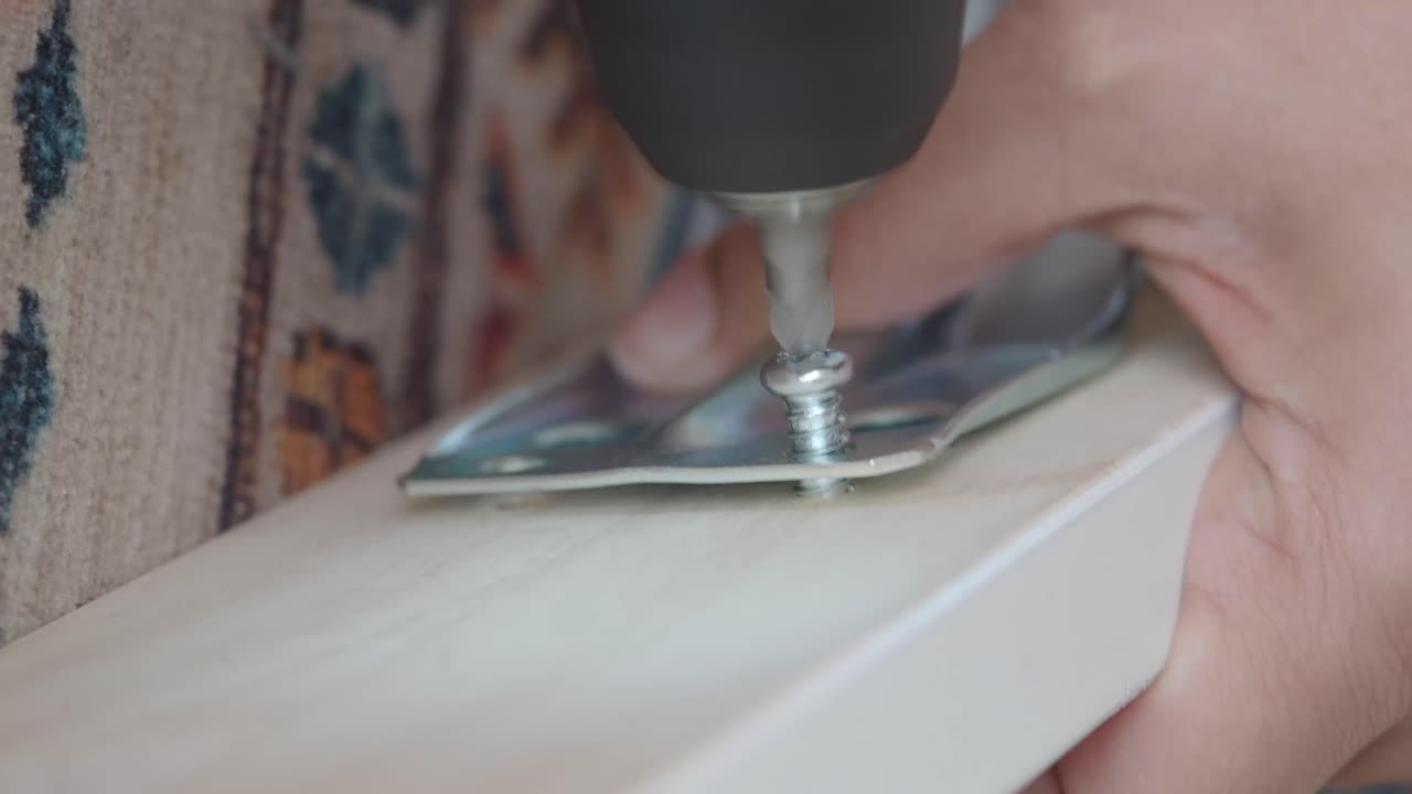 Primo piano di una mano che usa un trapano elettrico per avvitare una staffa metallica su del legno.