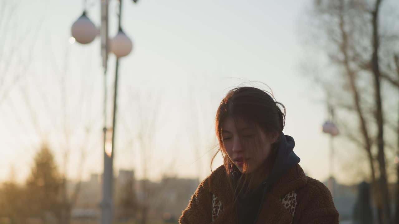 한 우울한 소녀가 일몰에 공원을 걸어가며 머리카락이 얼굴을 부분적으로 고, 갈색 코트와 후디를 입고, 그녀는 생각에 빠진 것처럼 보이며, 부드럽고 희미한 빛으로