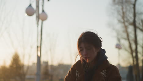 a depressed girl walking through a park at sunset with her hair partially covering her face. dressed in a brown coat and hoodie, she appears lost in thought, with the soft, fading light