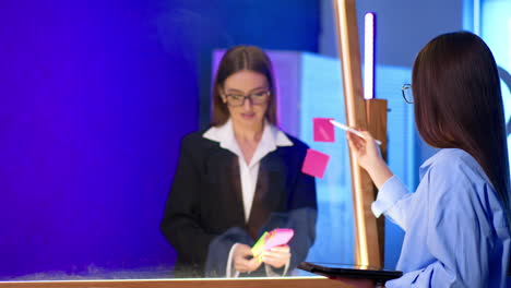brunette wearing black jacket taking stickers off the glass board. woman at foreground uses a tablet.