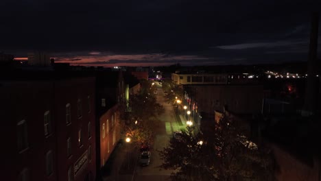 warm lighting lantern along main street of american town with red brick houses at night. rising drone wide shot. sunset at horizon at dusk. parking cars on road.