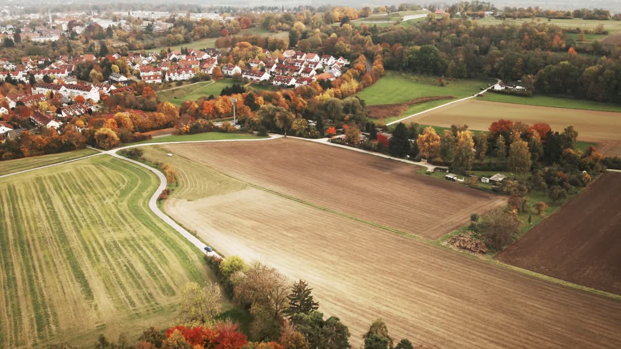 Camera glides forward over dense golden treetops and patchwork fields, approaching a picturesque German village nestled in seasonal colors