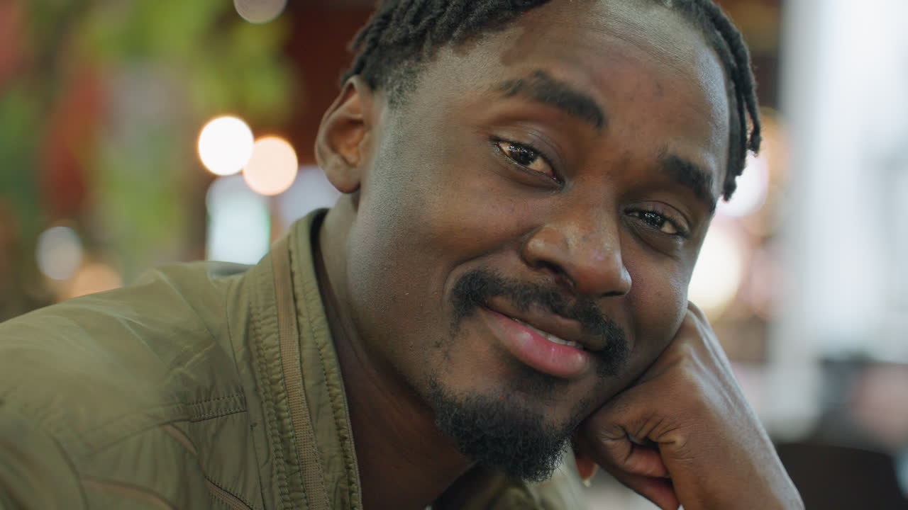 Close up of remote worker posing with calm smile eyes closed resting hand under chin in relaxed thoughtful mood, wearing green jacket indoors with blurred bokeh lights and soft warm background