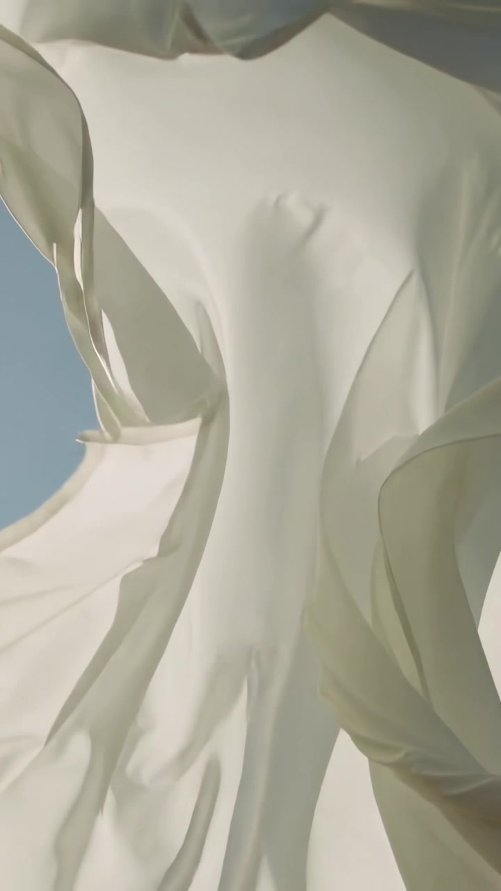 Video verticale: Folata di vento che solleva un tessuto bianco trasparente, rivelando pannelli a strati e cuciture contro il cielo blu.