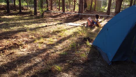 mujer joven descansando leyendo un libro en el campamento cerca de la tienda azul en el bosque de pinos silvestres