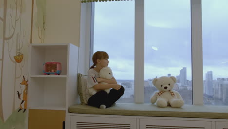 girl sitting on a window seat with stuffed animals, looking out at a city view.