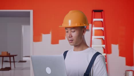 un pintor asiático que usa un casco de seguridad usando una computadora portátil mientras está de pie con una pared de pintura en la habitación