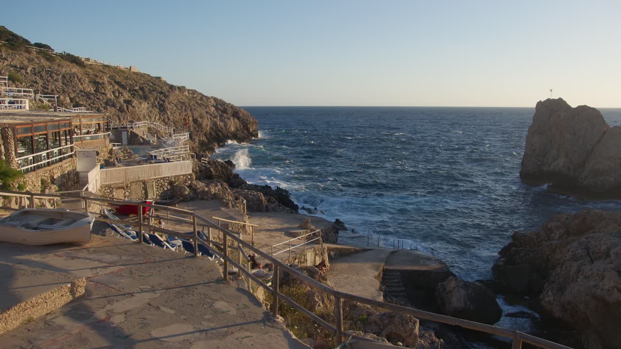 Wide shot showing waves crashing on rocks and part of a beach club during sunset at Faro di Punta Carena
