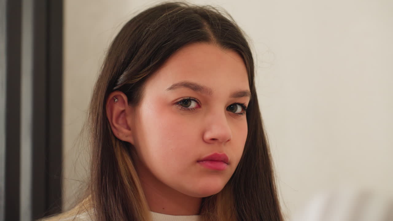 Portrait view of beautiful girl seated indoors, long natural hair and ear piercing visible, soft window light, wooden divider backdrop, subtle pout, neutral wall space framing youthful expression