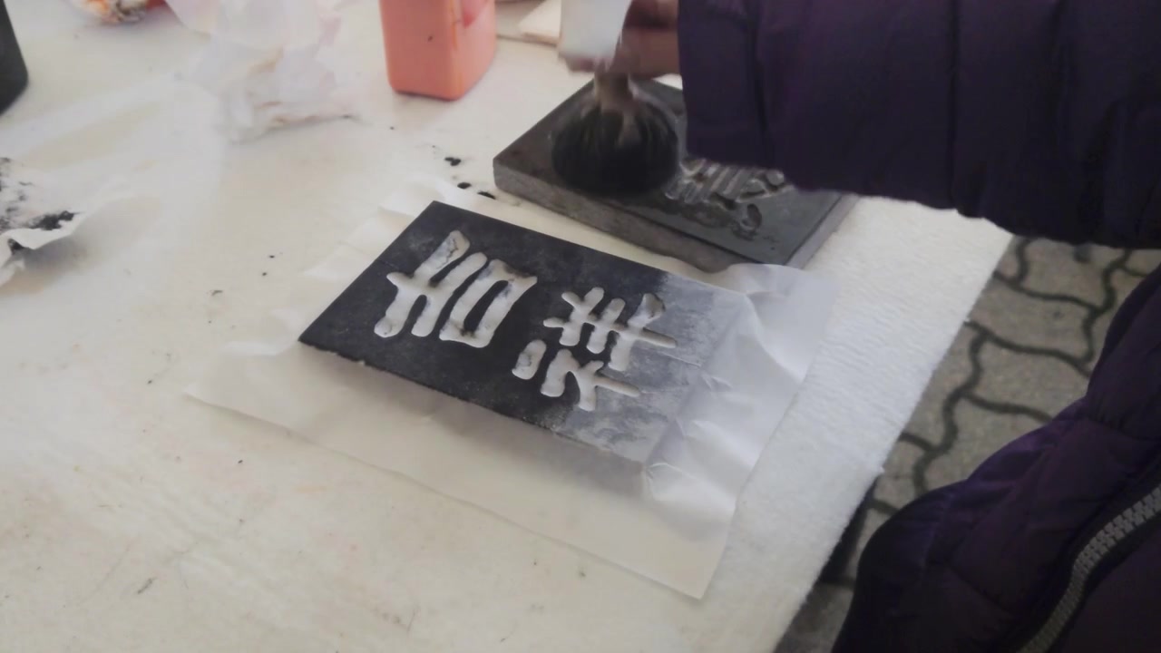 Outdoor static close up shot captures a participant using a round ink dabber tool to apply black ink evenly onto a carved wooden calligraphy printing matrix at a street festival workshop booth.