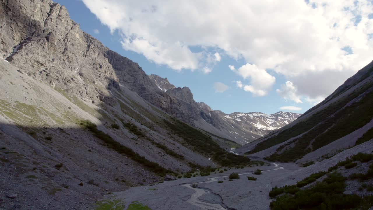 Aerial drone footage slowly descending into a glacial valley surrounded by a steep and dramatic mountain landscape with residual patches of snow and an alpine river in Switzerland
