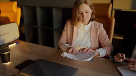 Turn-on-a-confident-middle-aged-blonde-girl-in-a-pink-business-suit-passes-papers-to-her-colleague-and-talks-about-her-plans-and-ideas-while-working-in-the-office-at-the-table