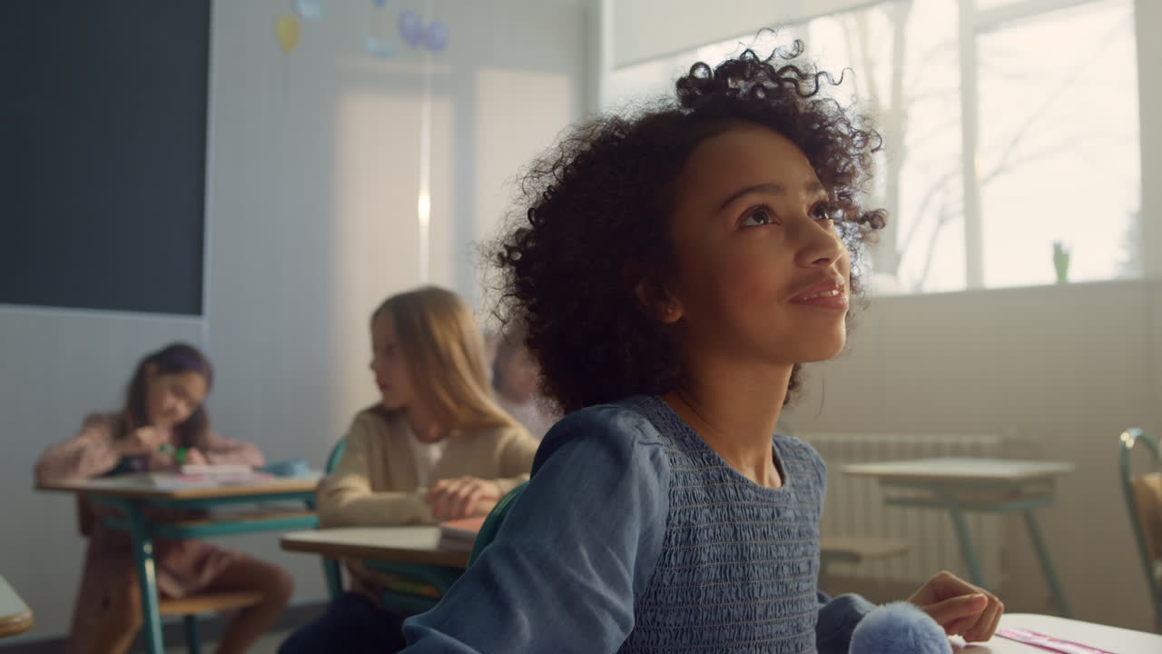 Positive afro girl learning in classroom. Cheerful student talking with teacher