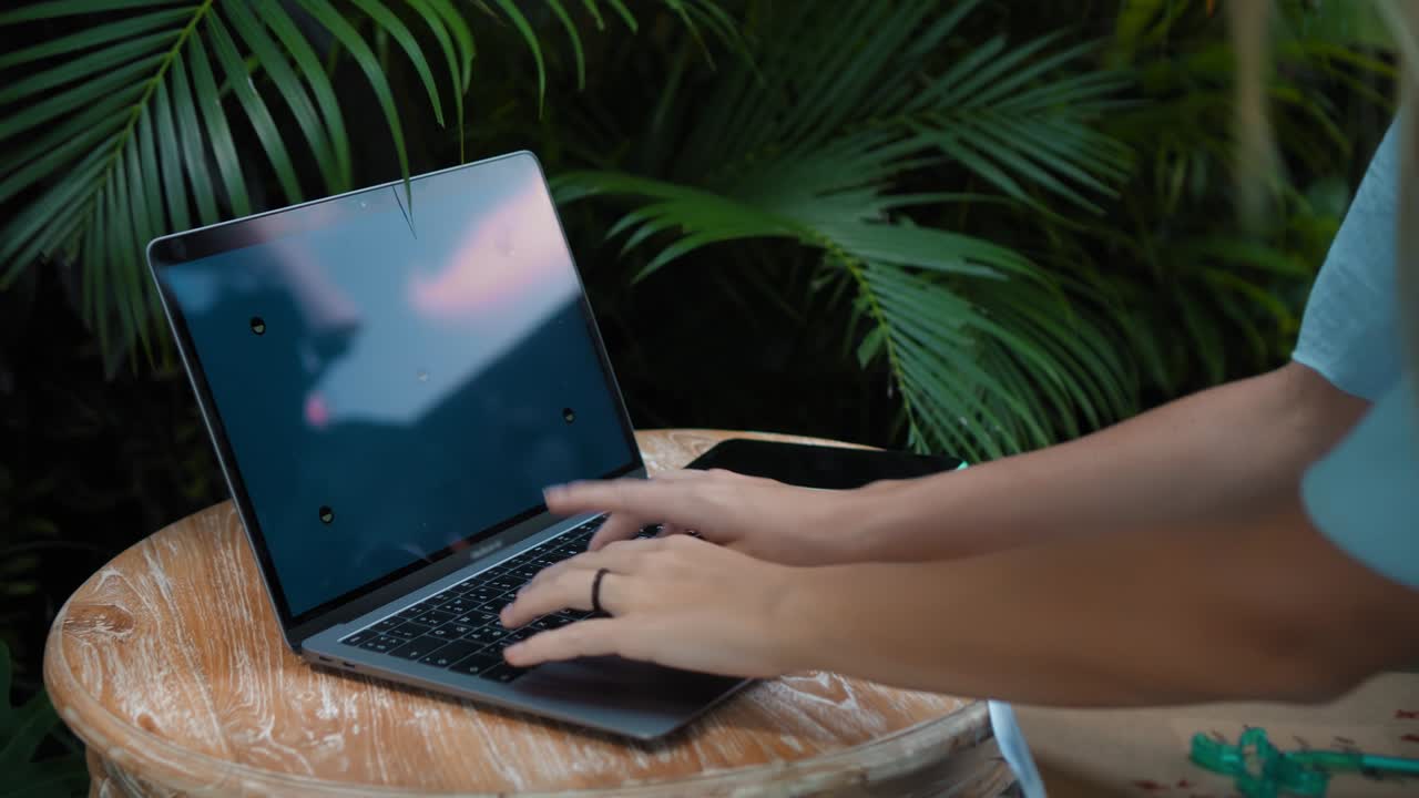 Una giovane europea in abito blu che lavora da remoto online su un laptop con schermo blu nel cortile con piante verdi