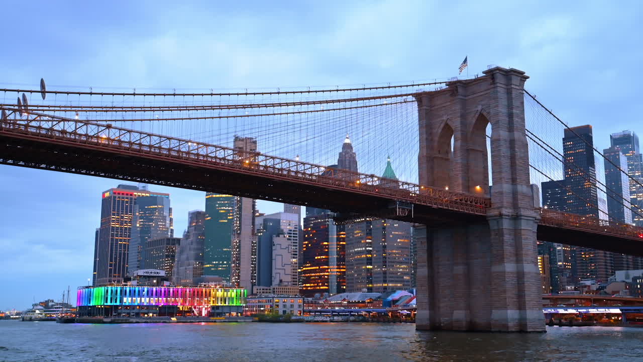 Approaching the Brooklyn Bridge by the river. Luminous New York skyline at backdrop. Low angle view