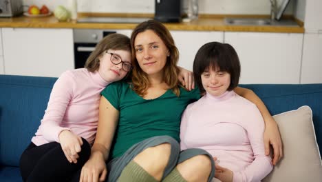 Portrait-of-two-girls-with-down-syndrome-in-sitting-on-a-couch-together-embracing-with-their-mom,-close-up