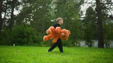 kid dressed in black walking through lush grassy field clutching large plush teddy bear, surrounded by tall trees partially hiding distant building in calm outdoor setting under natural daylight