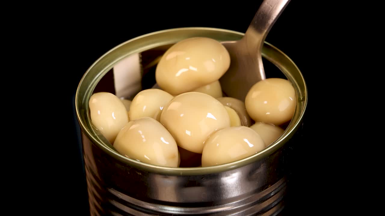 A metal spoon lifts whole champignon mushrooms from an open canned food tin against a black background, with bright, even studio lighting and close-up perspective
