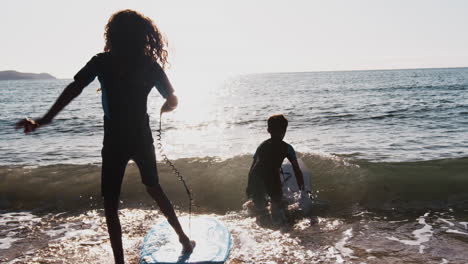 dos niños con siluetas que llevan trajes de buceo jugando en el mar con bodyboards en vacaciones de playa de verano