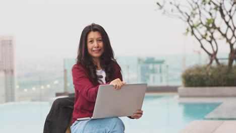 mujer de negocios asiática confiada en suéter rojo posando en un rascacielos con computadora portátil. feliz joven profesional, líder de negocios, jefe o entrenador corporativo mirando a la cámara, vista de la ciudad y sonrisa.