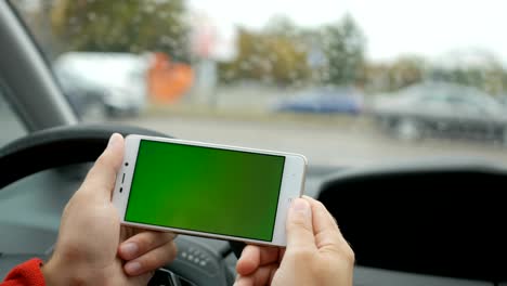 mano sosteniendo teléfono inteligente con pantalla verde en blanco en el coche para la dirección, masaje, ubicación, negocio. vida de la ciudad. hombre se sienta en el coche y trabaja en el teléfono inteligente pantalla verde de cerca. llave de croma. día lluvioso.