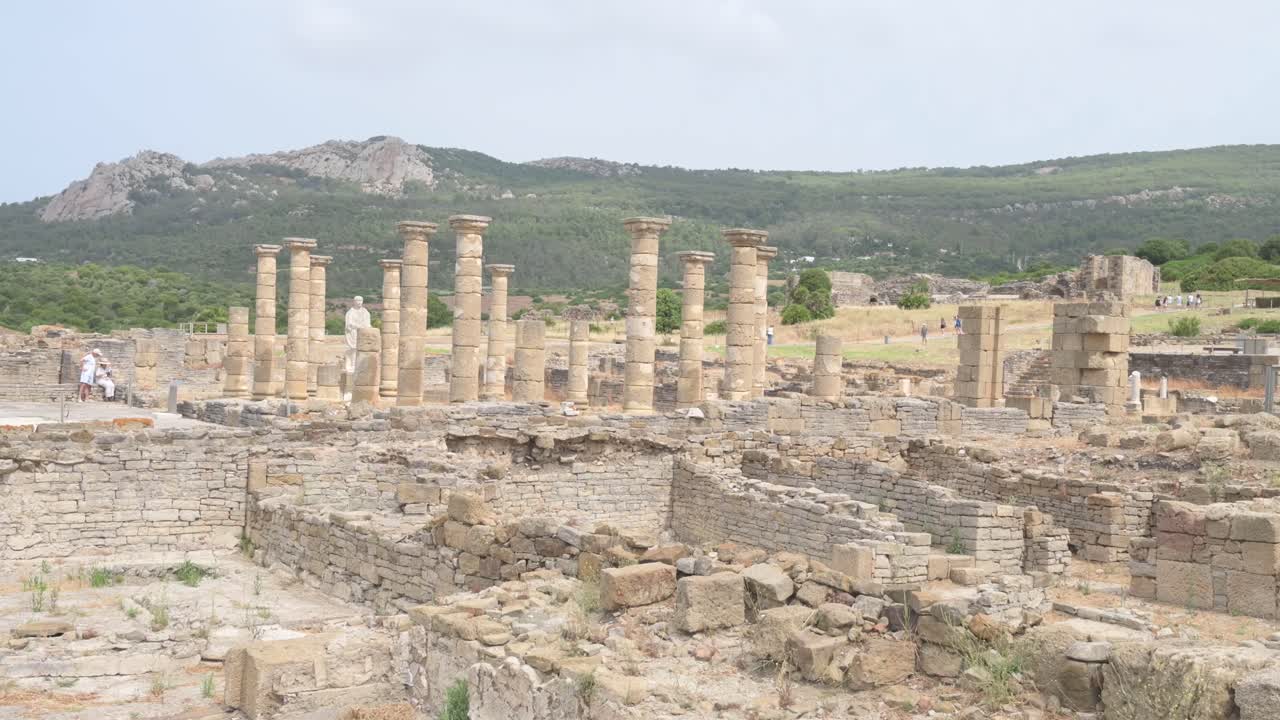 Wide shot of ancient Roman ruins of Baelo Claudia with scattered stone blocks and columns. Mountains in background