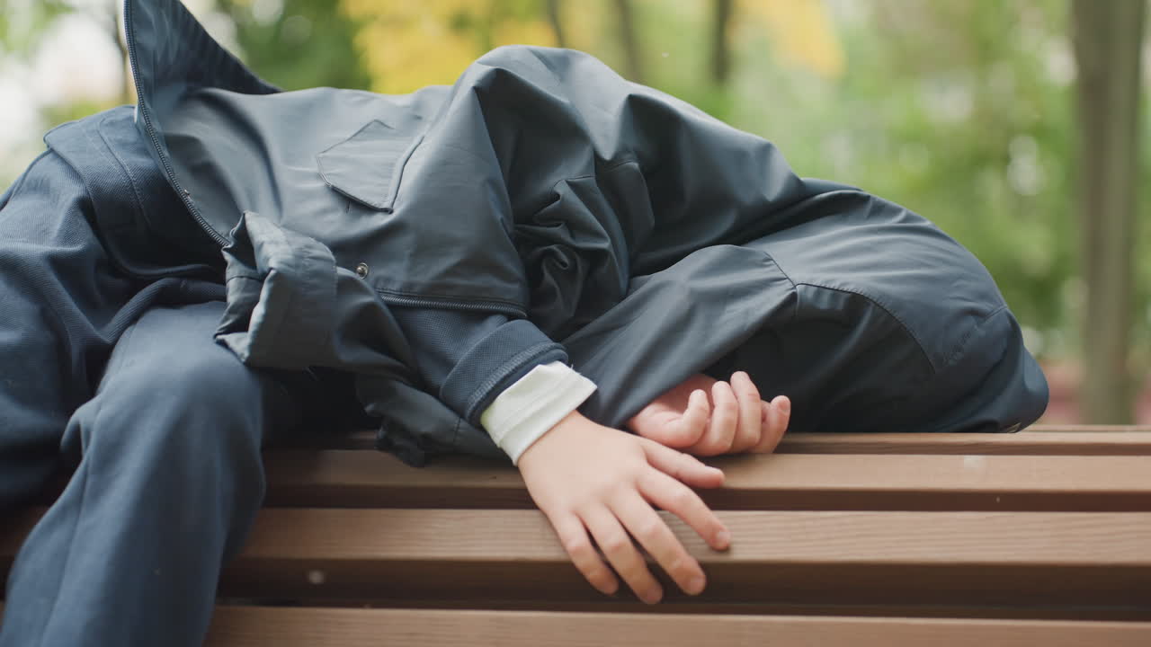 静かに休む学生。放課後、公園のベンチで眠る若者。木製の公園の座席で静かに眠る若い個人。秋の公園で、木製のスラットに手を置いて休む白人系の若者。