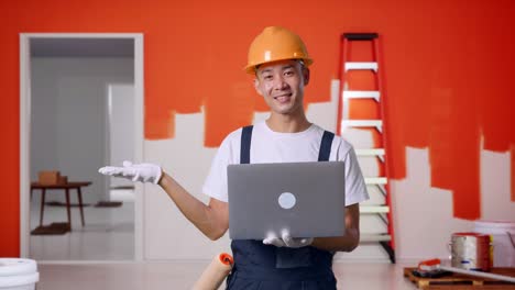 pintor asiático con casco de seguridad usando una computadora portátil y apuntando a un lado mientras está de pie con la pared de pintura en la habitación