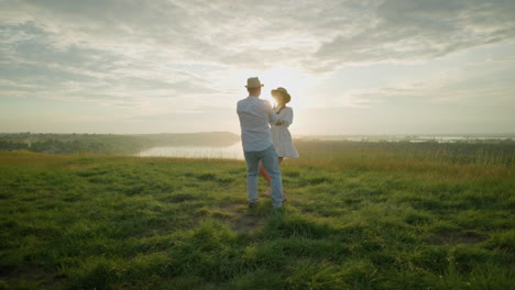 los amantes giran amorosamente en una colina cubierta de hierba al atardecer, al lado de un lago sereno. el hombre, vestido con una camisa blanca, sombrero y vaqueros, abraza a su esposa, que lleva un vestido blanco fluido, mientras disfrutan