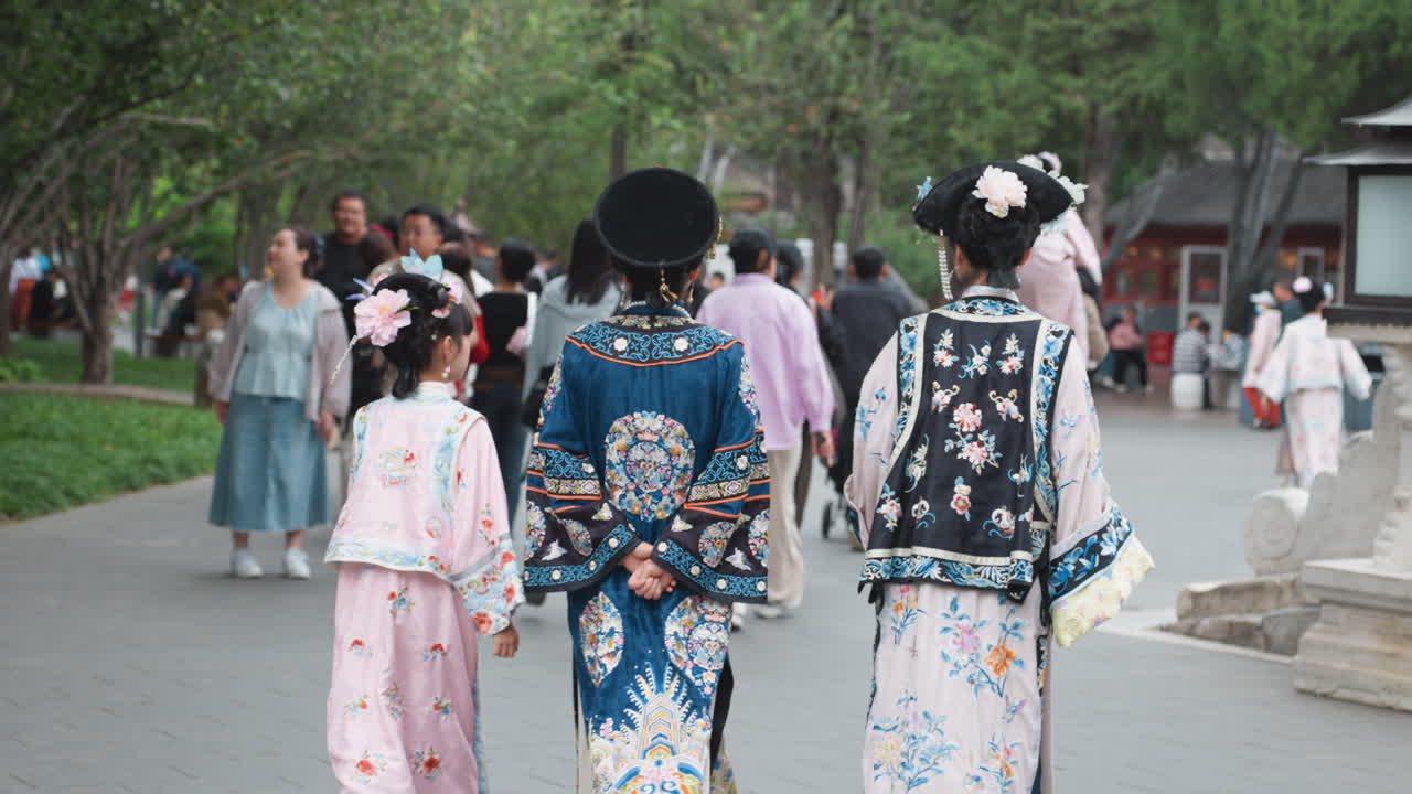 賑やかな人混みの中で、寺院の公園敷地を歩く漢服姿の三人の女性、刺繍の絹衣と花の簪を身につけ、石灯籠が並ぶ夏の午後、観光客が撮影する優雅な行列