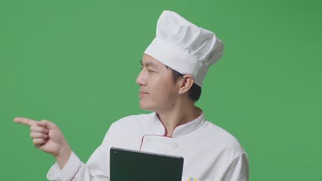 primer plano de un chef asiático usando una tableta y apuntando a un lado mientras está de pie en el estudio de fondo de la pantalla verde